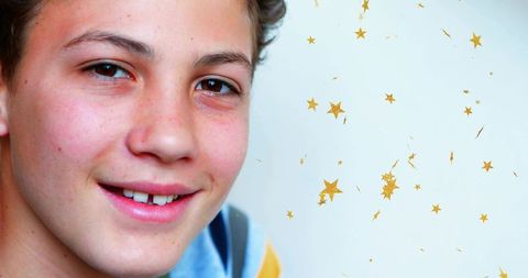Smiling boy wearing blue top with yellow stripe, freckles and gold star confetti on white backdrop