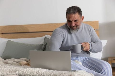 Senior man in pajamas working on laptop and sipping coffee on bed during relaxed morning