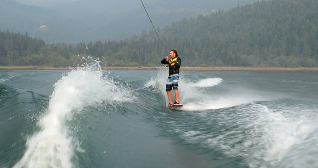 Man wakeboarding on calm lake surrounded by mountainous terrain. Suitable for themes related to watersports, outdoor recreation, adventure tourism, summer activities, and nature enjoyment. Perfect for promoting travel destinations, adventure sports equipment, and lifestyle blogs.