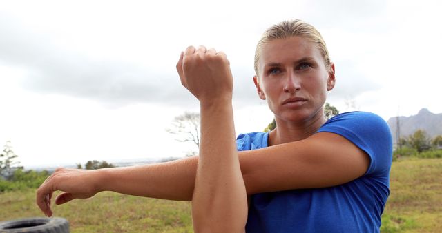 A young Caucasian woman is stretching her arm muscles outdoors, with copy space. Her focused expression and active posture suggest she is preparing for a workout or engaging in a fitness routine.