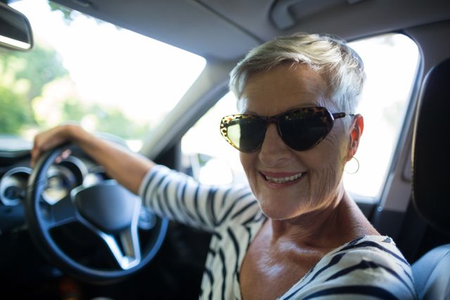 Senior woman confidently driving a car while wearing sunglasses and smiling. Ideal for use in advertisements promoting senior independence, lifestyle blogs, travel agencies, and automotive services targeting elderly customers.