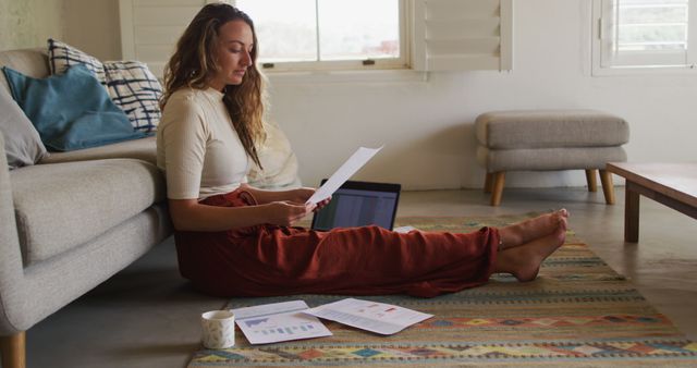 Caucasian woman working from home, sitting on living room floor with laptop, coffee and paperwork. working at home in isolation during quarantine lockdown.