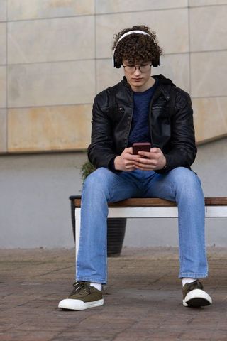 Teen sitting on bench using smartphone wearing headphones and leather jacket
