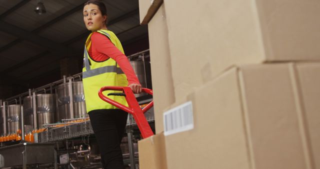 Female warehouse worker wearing safety vest moving large boxes with hand pallet truck in industrial setting. Ideal for content on logistics, physical labor, warehouse management, and supply chain topics.