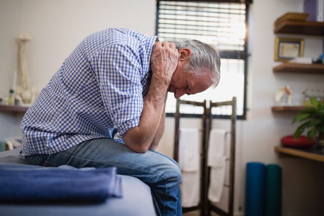 Side view of senior man sitting with neck pain on bed at hospital ward