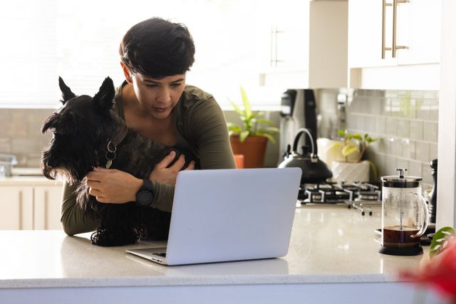 Caucasian mid adult lesbian woman embracing scottish terrier while looking at laptop in kitchen. Copy space, technology, unaltered, dog, pet, care, love, togetherness, friendship, lifestyle and home.