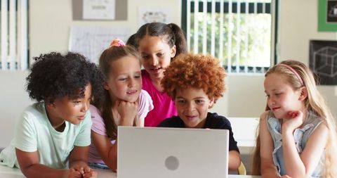 Diverse children collaborating on laptop while learning together in bright classroom