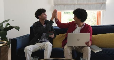 African American friends high-fiving on couch using laptop and tablet, remote work vibe