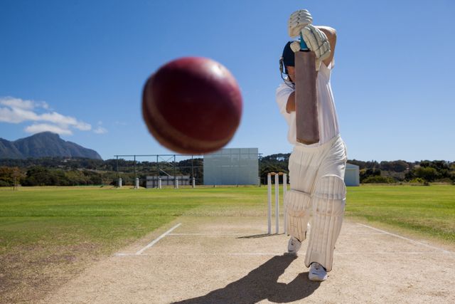 Batsman playing cricket, wearing white uniform, hitting ball on pitch with clear blue sky and mountains in background. Perfect for use in sports promotions, cricket training materials, outdoor activity advertisements, and athletic event posters.