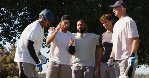 Diverse male cricket team gathering and discussing strategy on sunny field with bat