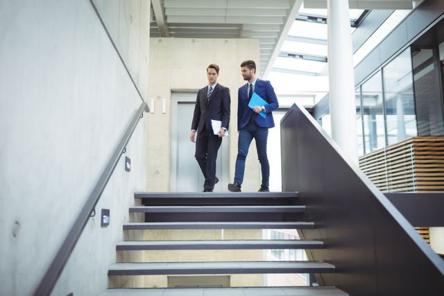 Two businessmen are walking down the stairs in a modern office building, engaged in a discussion. Both are dressed in professional business attire, holding documents. This image can be used for corporate presentations, business websites, teamwork and collaboration themes, or office environment illustrations.