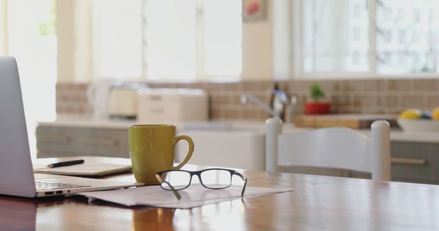 This image showcases a cozy home office setup, with a yellow mug, glasses, and an open laptop on a wooden desk. A bright kitchen is visible in the background, creating a pleasing and productive work environment. Perfect for illustrating concepts of remote work, home productivity, and modern lifestyle aesthetics. Useful for blog posts, articles, and brochures centered around work-from-home setups or home interior designs.