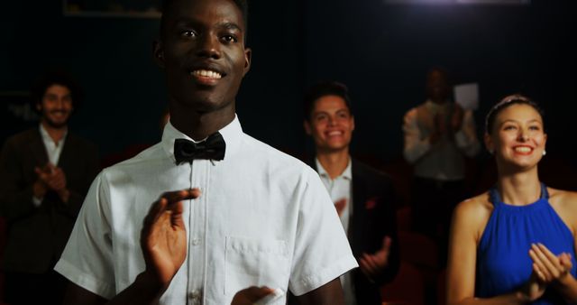 A young African American waiter in a bow tie stands in the foreground, with a diverse group of people applauding in the background, with copy space. His confident smile and the applause suggest a celebration or a successful event.