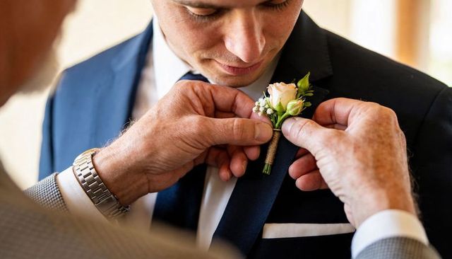 Father pinning blush rose boutonniere on groom in navy suit closeup