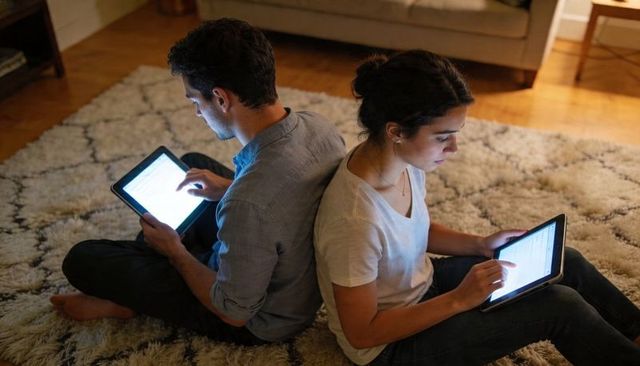 Young couple sitting back-to-back using tablets on cozy plush rug under warm evening light