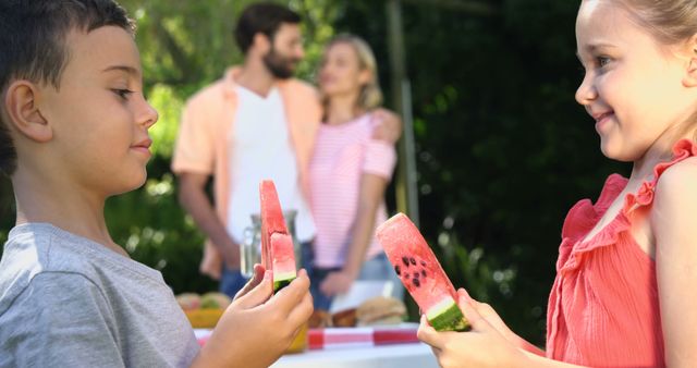 A boy and a girl enjoy eating watermelon at an outdoor gathering, with copy space. In the background, a man and a woman engage in a conversation, adding to the relaxed, social atmosphere.