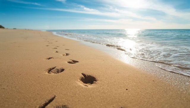 Footprints Leading Along Sunny Beach Toward Horizon with Gentle Waves and Sun Glare