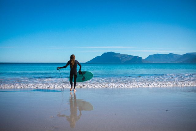 Surfer standing on sandy beach holding surfboard, looking at ocean with mountains in background. Ideal for promoting beach vacations, water sports activities, outdoor adventures, and active lifestyle content.