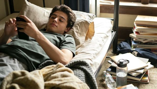 Male student lying on bed scrolling smartphone amid books, notebooks and water bottle