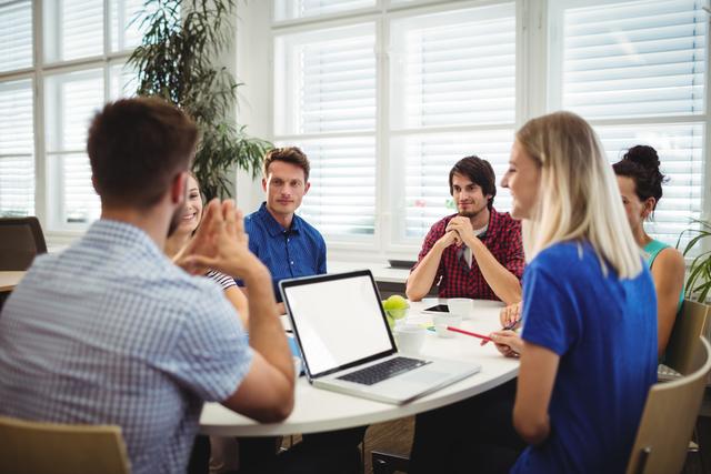 Young professionals around a table discussing ideas in a modern office environment. Ideal for articles about teamwork, business strategy sessions, corporate culture, or productivity tips in a collaborative workspace.