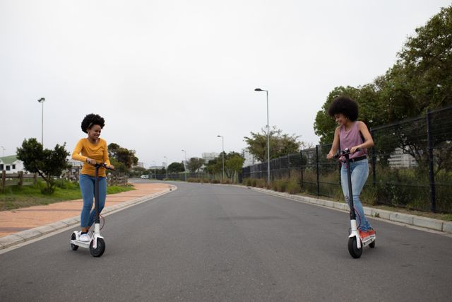 Front view of two happy biracial twin sisters enjoying free time together in a city, riding electric scooters in an urban park.