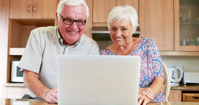 Elderly couple joyfully using laptop in contemporary kitchen. Perfect for illustrating themes of seniors embracing technology, retirement activities, lifelong learning, internet usage by the elderly, and modern kitchen interiors.