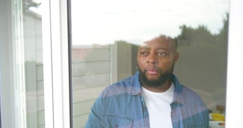 African man standing by sliding glass door looking contemplative through window at backyard