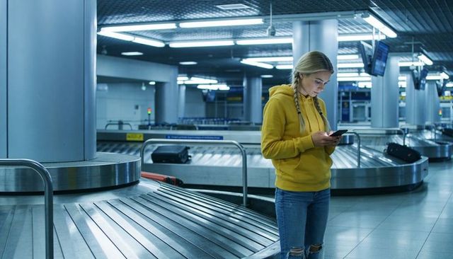 Young woman checking smartphone at airport baggage carousel in yellow hoodie
