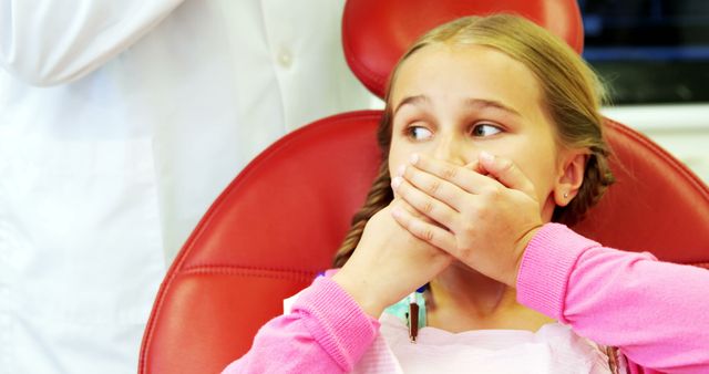 This visual depicts a young girl in a dental chair, appearing anxious while covering her mouth with her hands. The setting suggests a dental clinic and highlights emotions such as fear and anxiety associated with dental visits. Useful for illustrating concepts in pediatric dentistry, dental phobia, child patient care, and healthcare services.