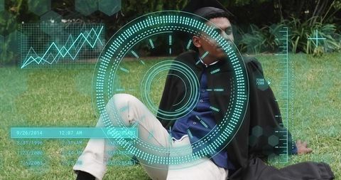 Male graduate relaxing on campus lawn wearing mortarboard and gown with futuristic hud