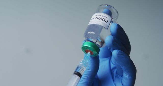 Close-up of a hand wearing a blue glove holding a vial labeled 'COVID-19' and inserting a syringe. This image conveys concepts of vaccination, medical science, and efforts to fight the COVID-19 pandemic. Perfect for articles, news stories, medical presentations, and educational materials related to vaccines, healthcare, and the pandemic response.
