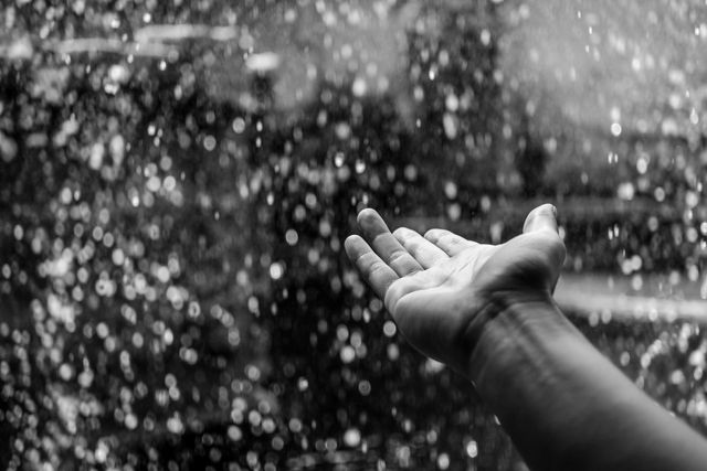 This image shows a hand reaching out beneath falling water droplets in a rainstorm, captured in black and white. The focus on the extended hand and the sparkling droplets creates a sense of connection with nature and the elements. Ideal for use in articles or promotions about rain, weather, human connection with nature, and mindfulness practices. It can also be used in artistic and inspirational contexts for galleries, blogs, and social media posts.
