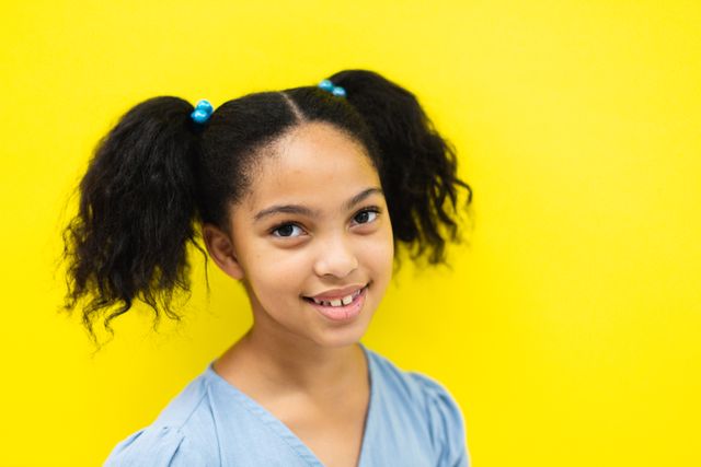 This image features a smiling biracial elementary schoolgirl with pigtails against a vibrant yellow background. Ideal for use in educational materials, advertisements for children's products, or campaigns promoting diversity and inclusion. The bright background and cheerful expression make it perfect for conveying positivity and youthful energy.