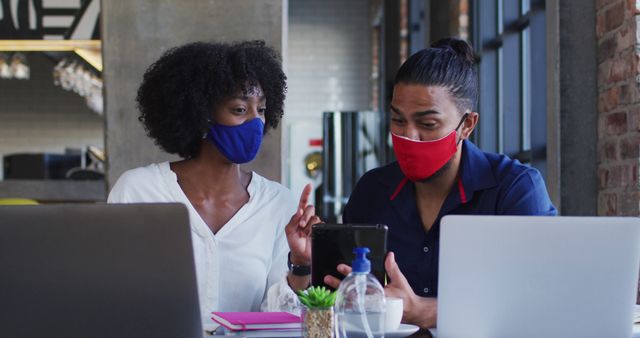 Two colleagues with masks are collaborating in a modern office space, discussing work on a digital tablet. Useful for depicting safe workplace practices, remote and office hybrid working environments, adapting to pandemic conditions, and teamwork in professional settings.