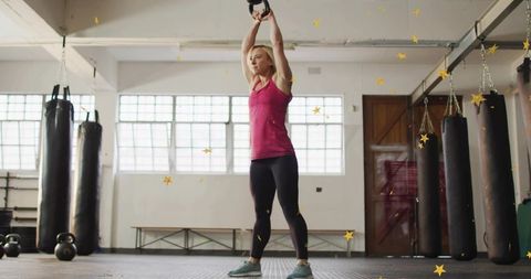 Woman lifting kettlebell overhead in industrial gym with punching bags and weights