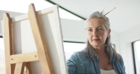 Mature woman painting at wooden easel in bright studio, natural light, creative focus