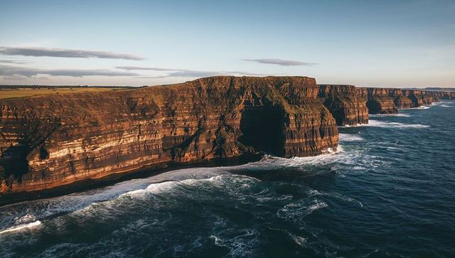 Layered coastal cliffs revealing sea cave and breaking waves along rugged shoreline