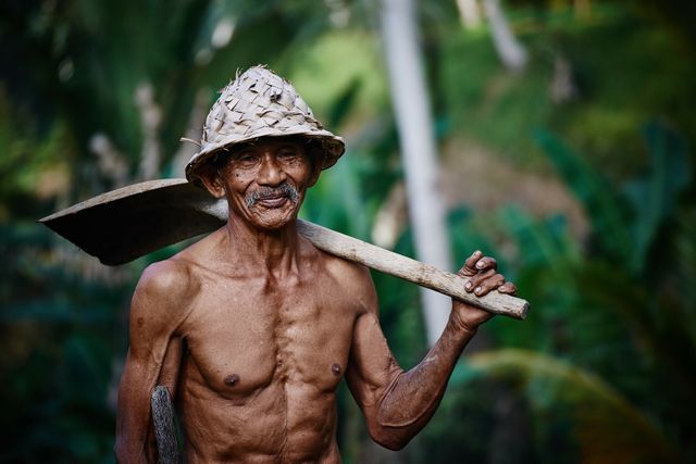 Elderly man in bamboo hat working in tropical fields, representing traditional farming and rural life. Useful for agricultural projects, cultural blogs, and articles discussing sustainable practices or elderly workforce in agriculture.
