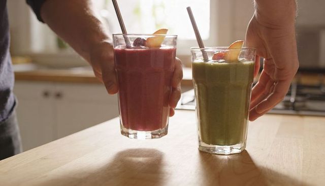 Two hands holding berry red and green smoothies with metal straws on wooden counter