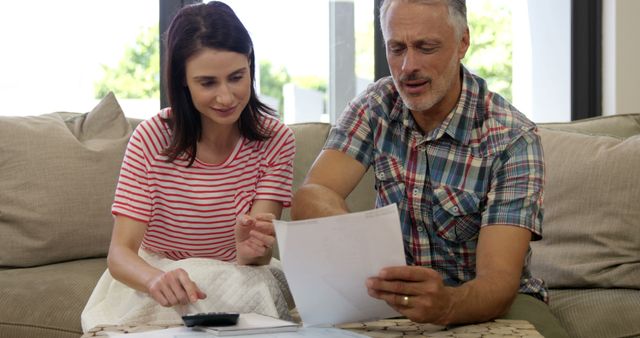 Couple sitting on a sofa reviewing financial documents with a calculator. They are working together, looking focused on managing their expenses or budget. This image is perfect for illustrating themes related to finances, family budgeting, financial planning, or household bills.
