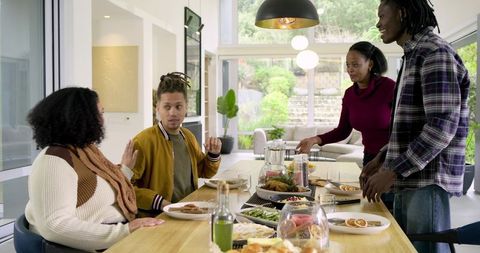 Multicultural friends gathering around wooden table sharing meal in sunlit modern home