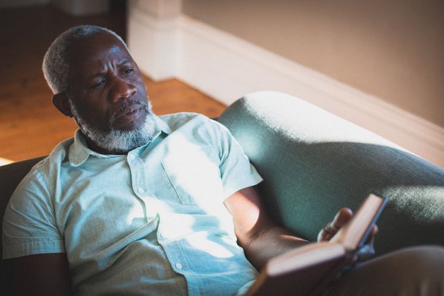 This image depicts a senior African American man lying on a couch while reading a book in a living room. The scene suggests relaxation and leisure, making it suitable for use in articles or advertisements related to home life, retirement, reading habits, or mental well-being during quarantine. The natural sunlight adds a warm and peaceful ambiance, ideal for promoting comfortable and serene home environments.