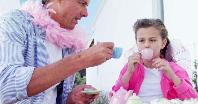 Father and daughter engaging in a playful outdoor tea party. Both are wearing fairy wings and boas, emphasizing a whimsical and imaginative bonding experience. Suitable for illustrating family bonding moments, childhood immersive play, and parent-child relationships in marketing materials, family blogs, and emotional storytelling campaigns.