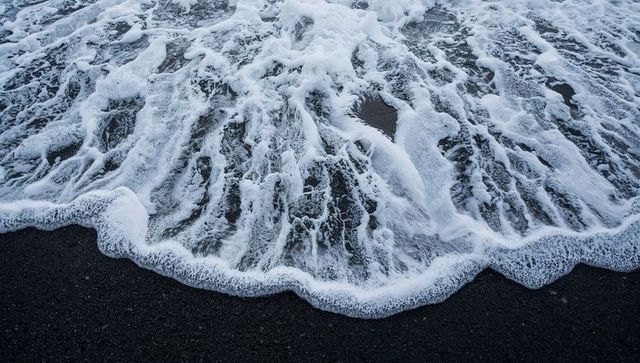 Close-up foamy ocean waves washing over black sand beach creating lace-like patterns