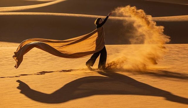Woman tossing sand on sunset dunes with flowing scarf and dramatic shadow