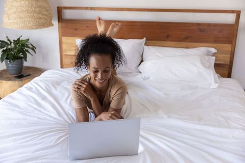 African american woman relaxing on white bed using laptop in modern minimalist bedroom