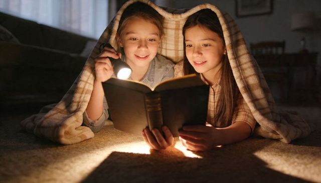 Two girls reading under plaid blanket by flashlight at night sharing cozy bedtime story