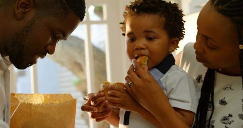 Parents feeding toddler homemade muffins by sunlit window enjoying cozy family breakfast