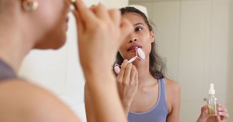 Asian woman using pink facial roller holding serum at vanity mirror for skincare routine