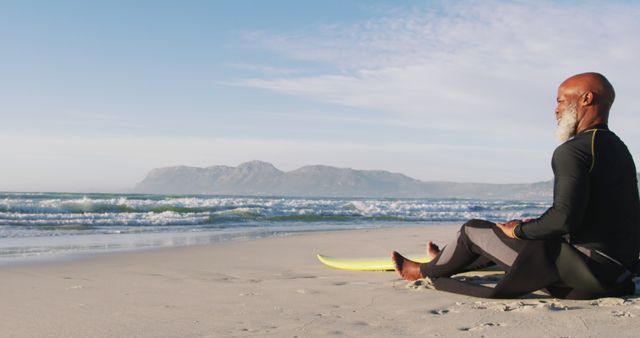 Senior man meditating on sandy beach with a yellow surfboard, gazing at the ocean horizon with distant mountains in the background. Ideal for themes focusing on wellness, relaxation, active aging, balance, mindfulness, retirement, and beach living.